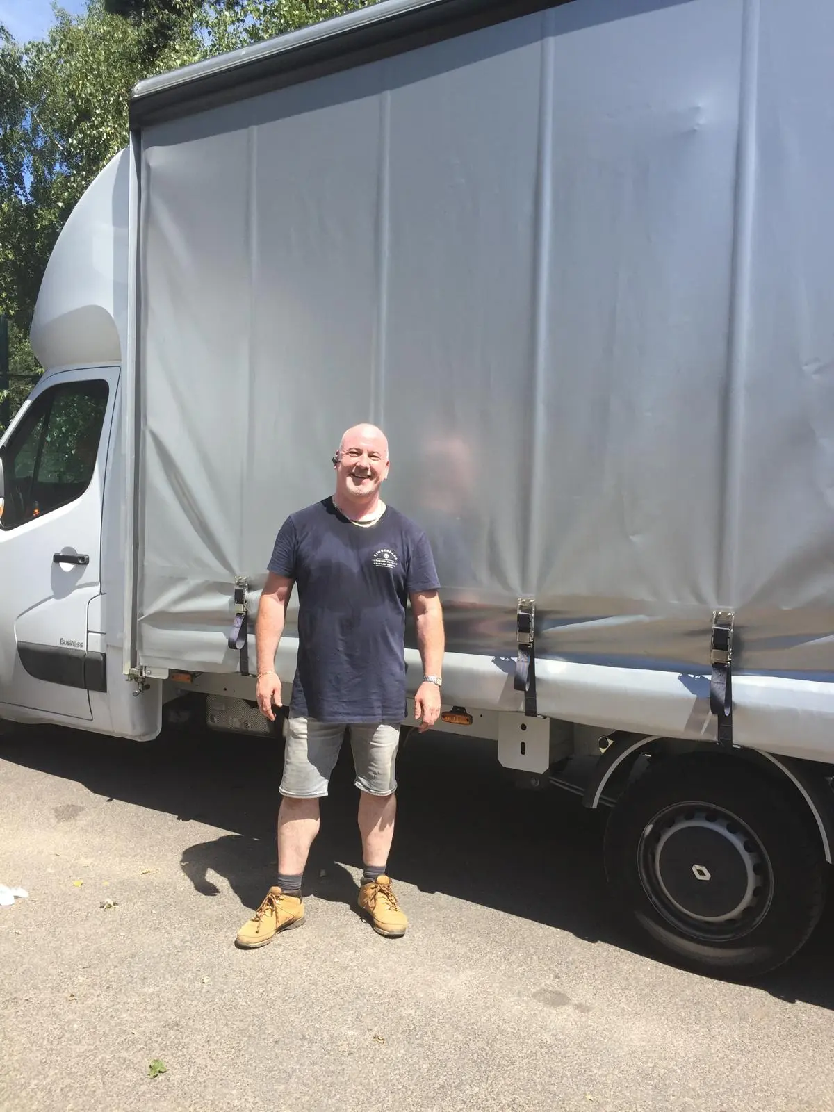 Simon Goulding stands smiling beside a large renault van. He's wearing a blue top, grey shorts, and yellow boots.