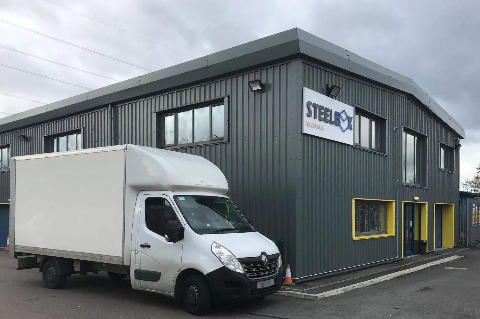 A photo taken outside the Steelbox building, which is two-storeys high, steel-clad, and has yellow window and door frames. A large white renault van is off to the left of the building.